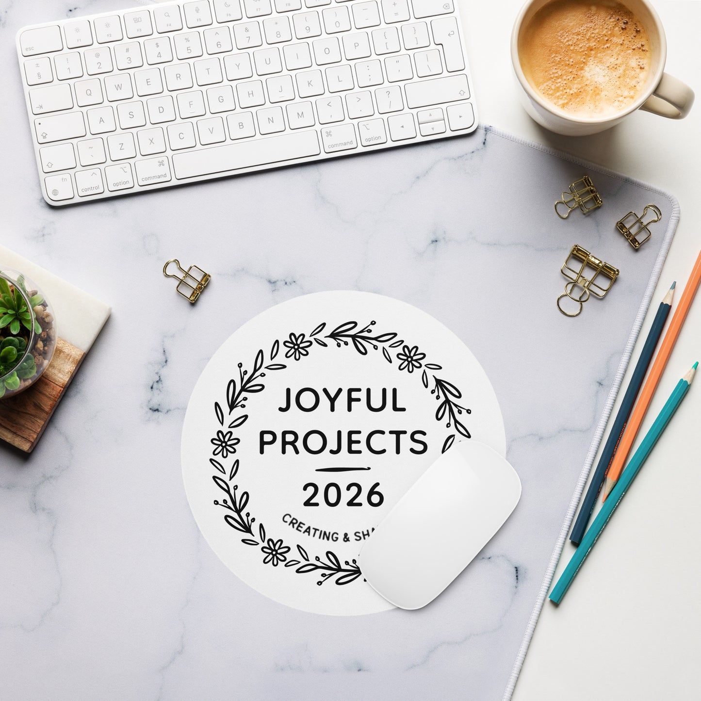 Minimal desk setup with keyboard, coffee, pencils, and a round Joyful Projects 2026 mouse pad on a marble surface.