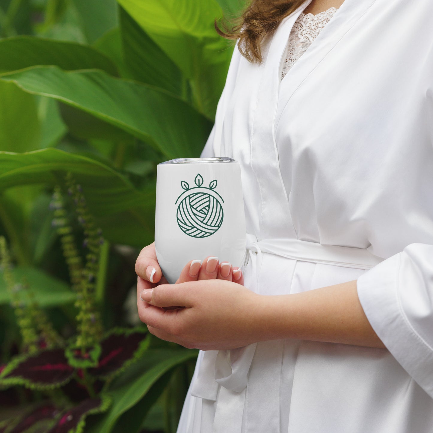 Person in a white robe holding the Stitch Sage 12 oz stainless steel tumbler, featuring the green yarn-ball-with-leaf-halo Maker Language™ icon, with lush green plants in the background.