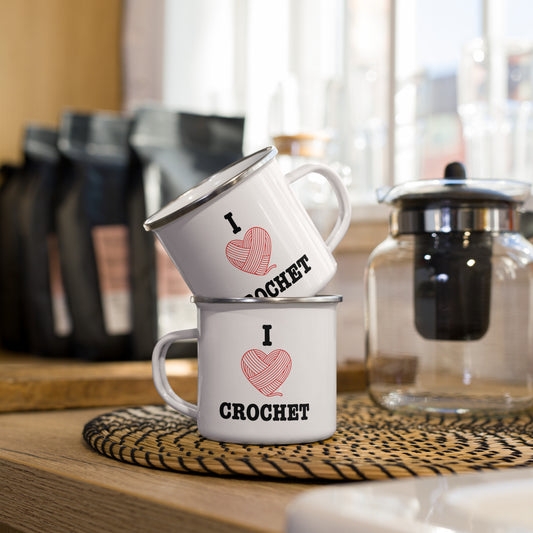 Two white I Heart Crochet enamel mugs stacked together on a cozy kitchen counter, showing the red yarn-heart design and CROCHET lettering. Warm lifestyle setting with coffee accessories in the background.